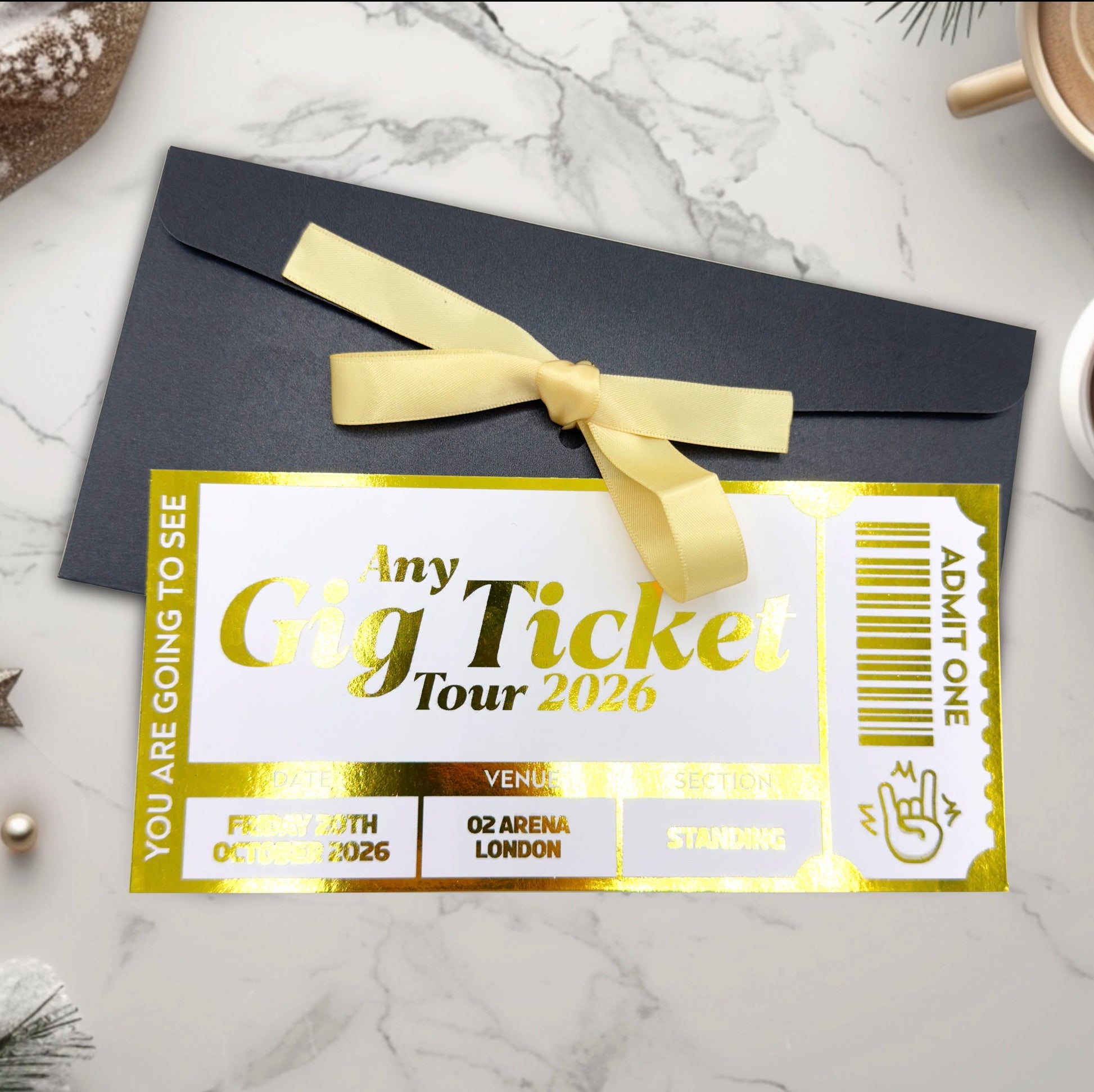 Personalised foil gig ticket with gold text and a yellow ribbon on a dark envelope against a marble background.